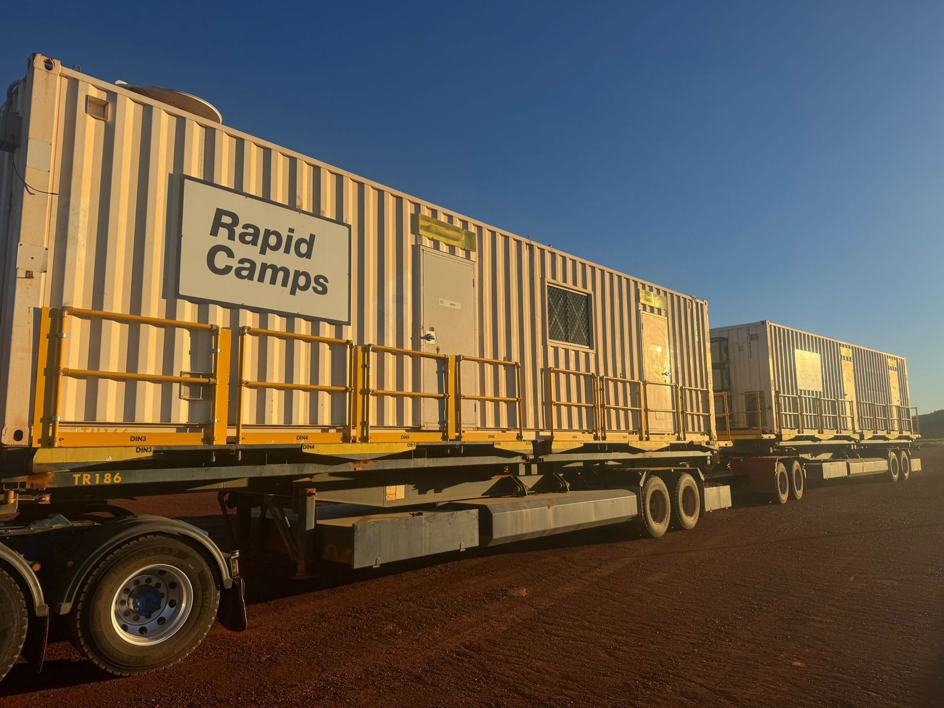 Two yellow cargo containers on flatbed trucks parked on a dirt road at sunset