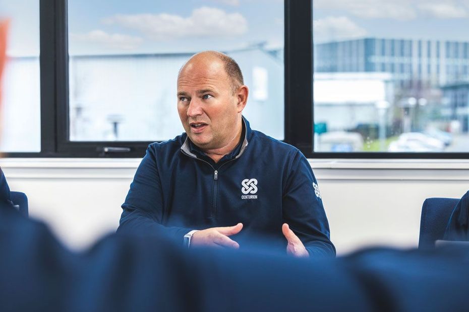 Man in blue jacket gesturing while speaking at a table, indoors near a window.