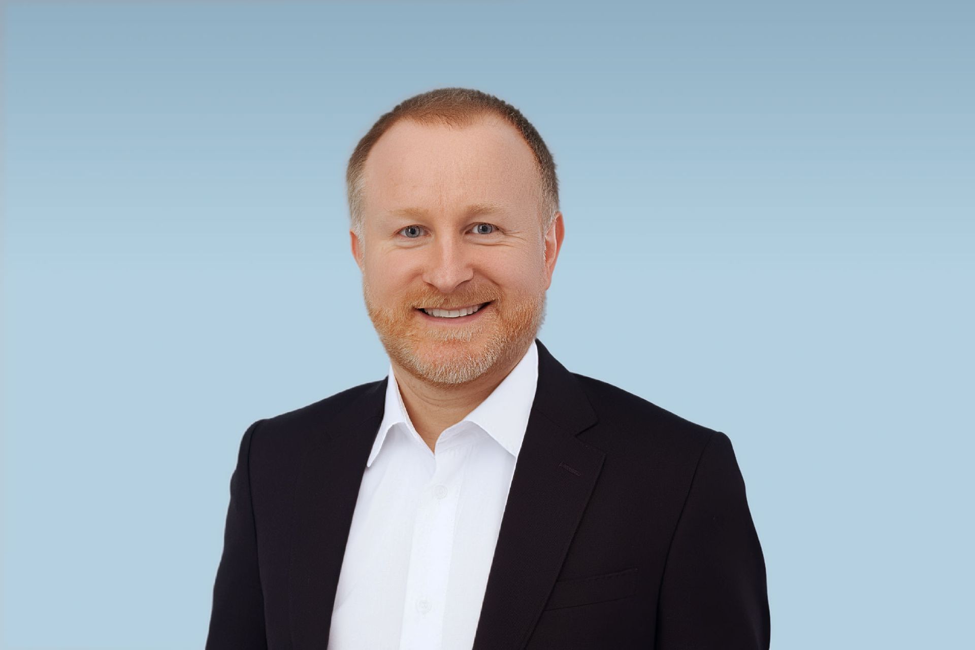 Man in a dark suit smiles, posed against a teal-green gradient background.