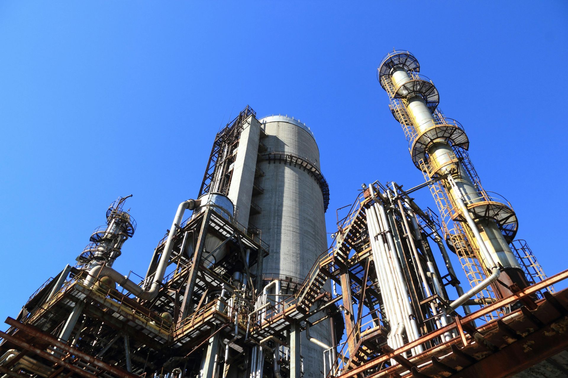 Industrial plant complex against a clear blue sky, featuring tall cylindrical structures and metal piping.
