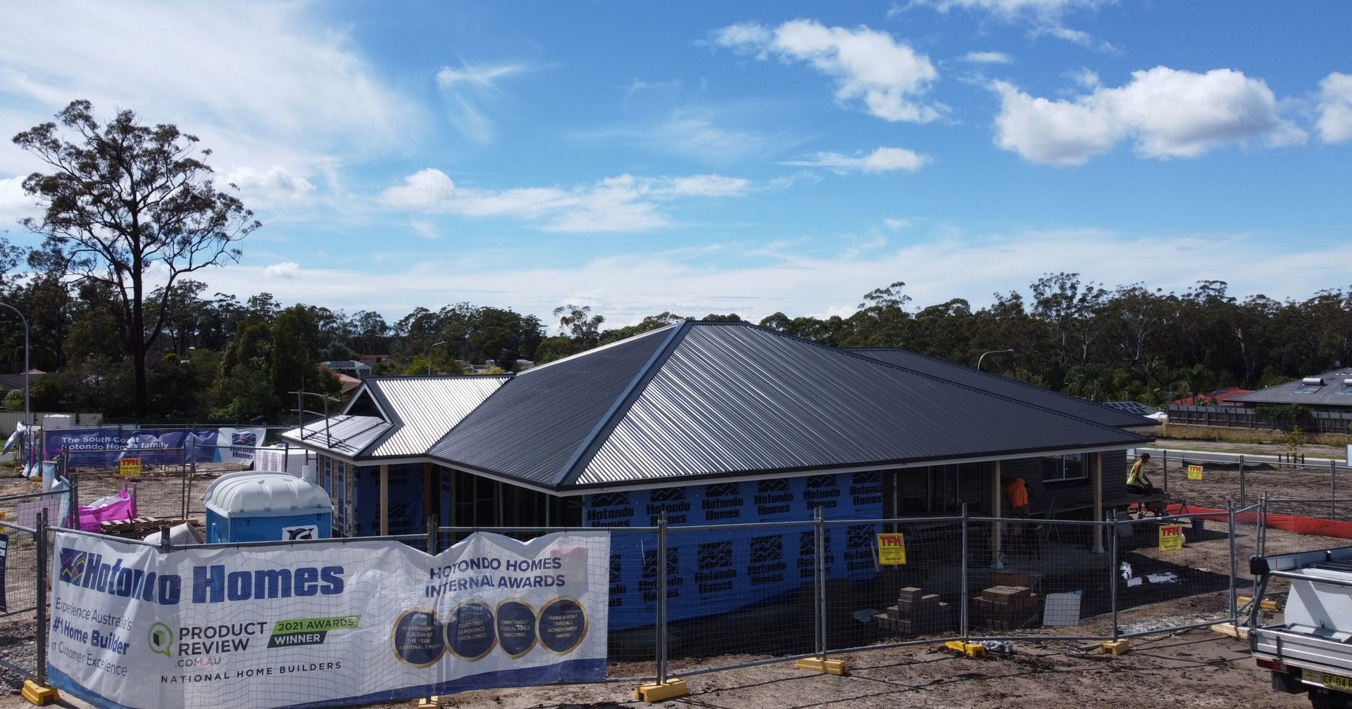 New house under construction; blue wrap, dark roof, sunny day. 