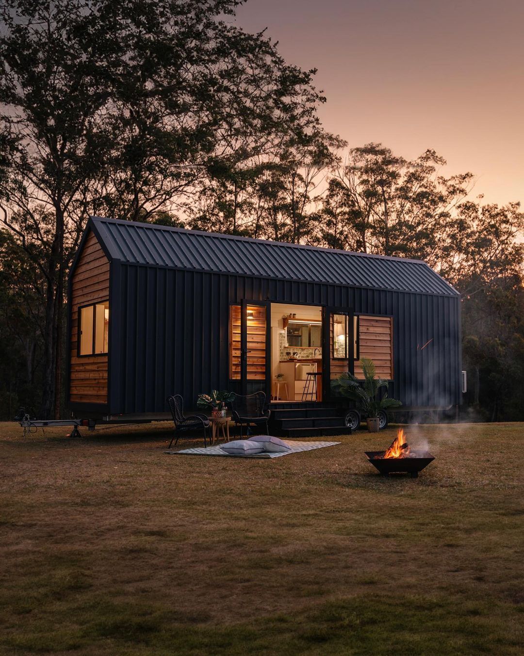 Tiny house with open door, fire pit, and outdoor seating at dusk.