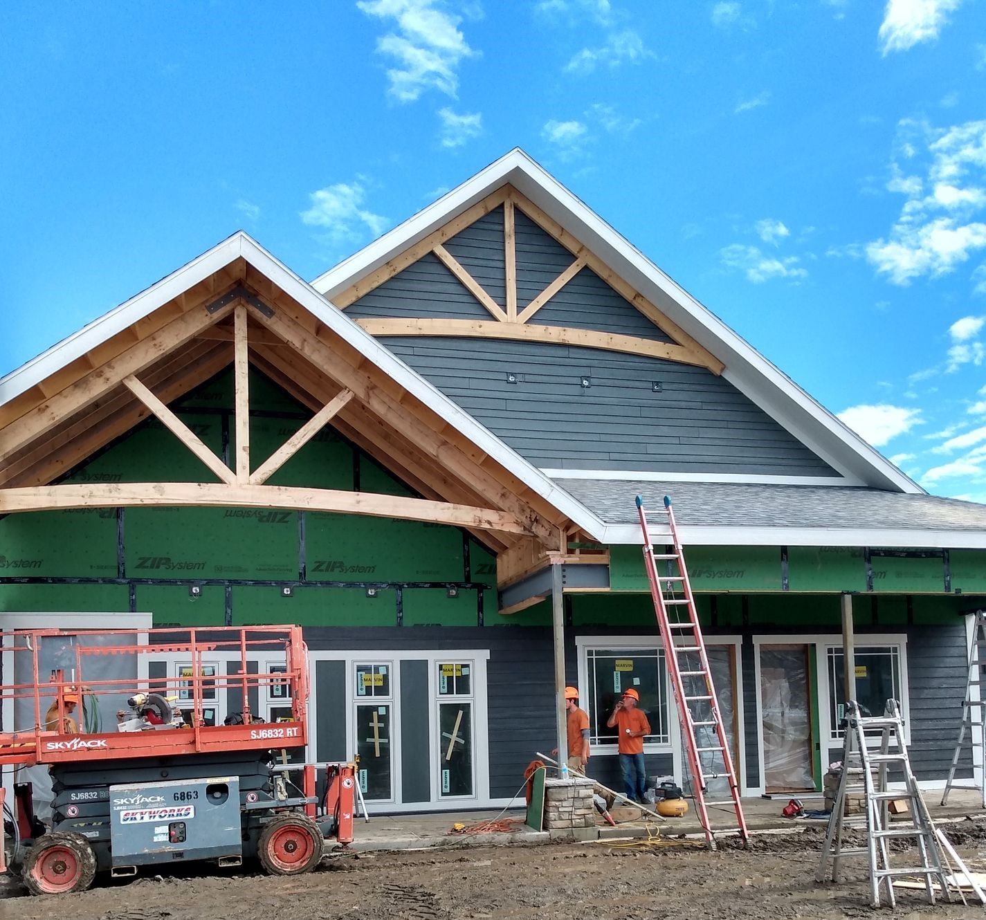 Construction of a house with exposed wood framing, dark gray siding, and a scissor lift; workers visible.
