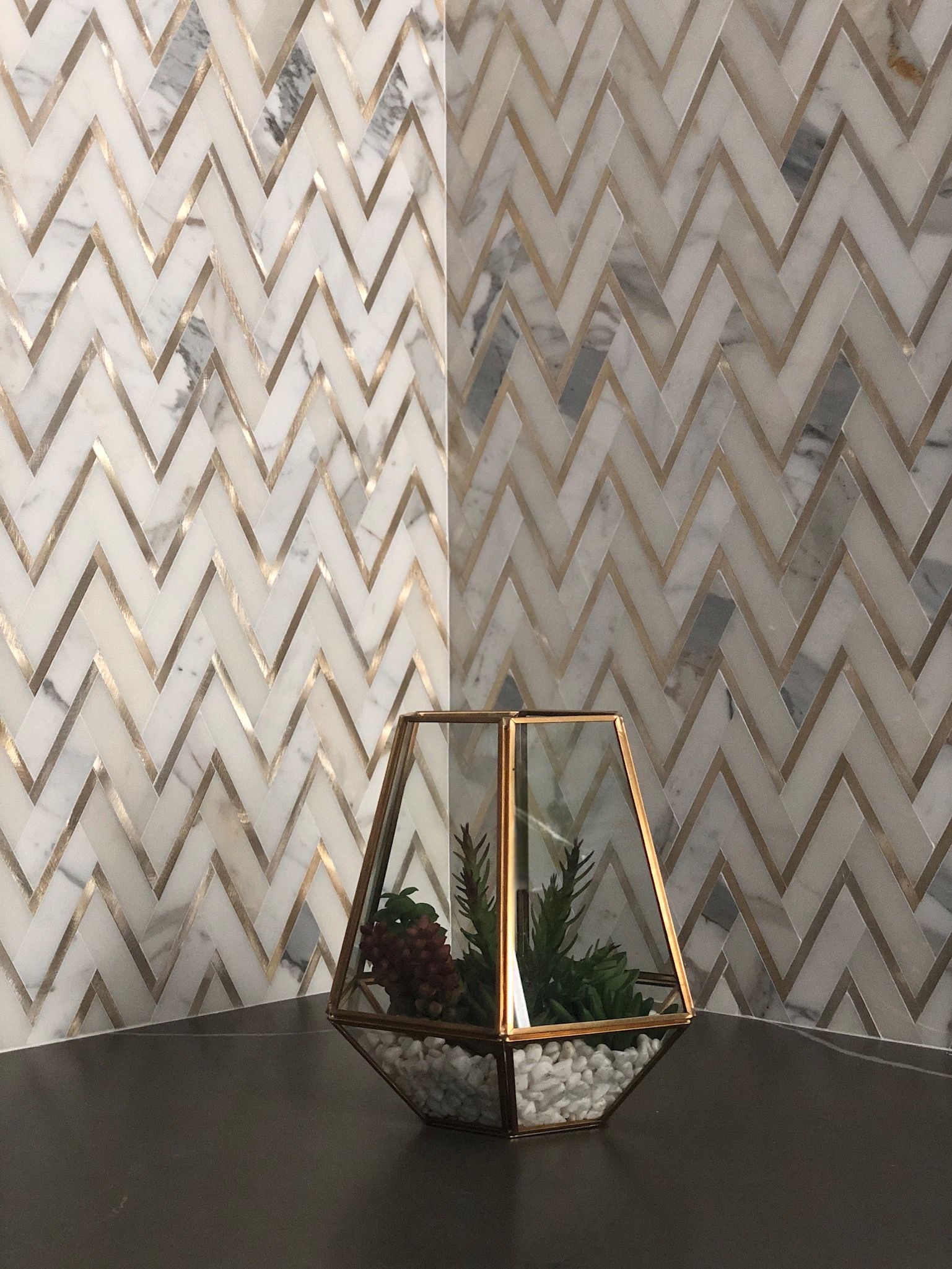 A glass vase with a plant in it is sitting on a table in front of a wall with a herringbone pattern.