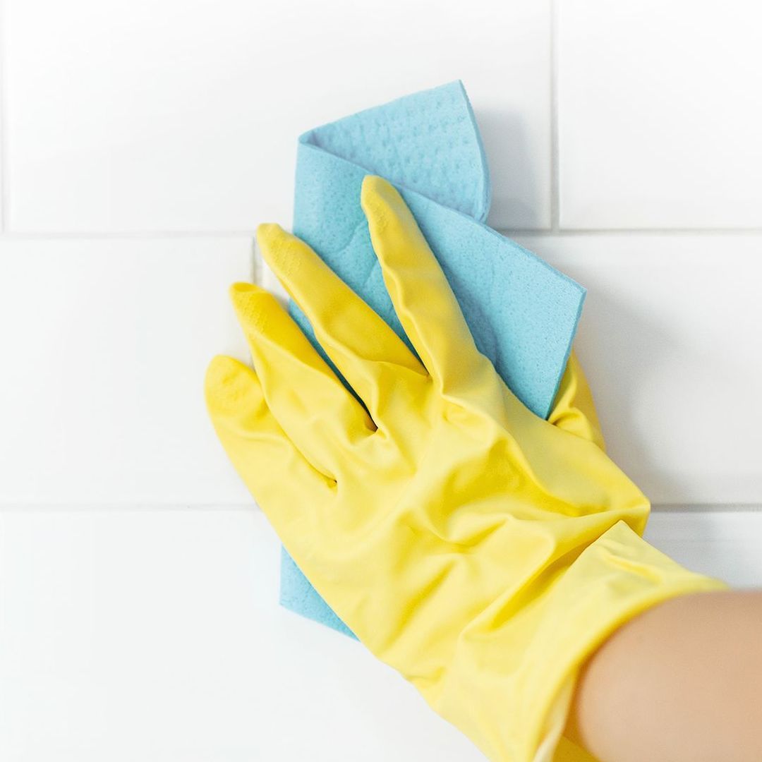 A person wearing yellow gloves is cleaning a tile wall with a blue cloth.