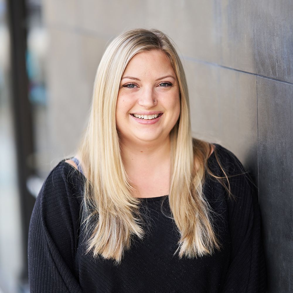 A woman with long blonde hair is leaning against a wall and smiling.