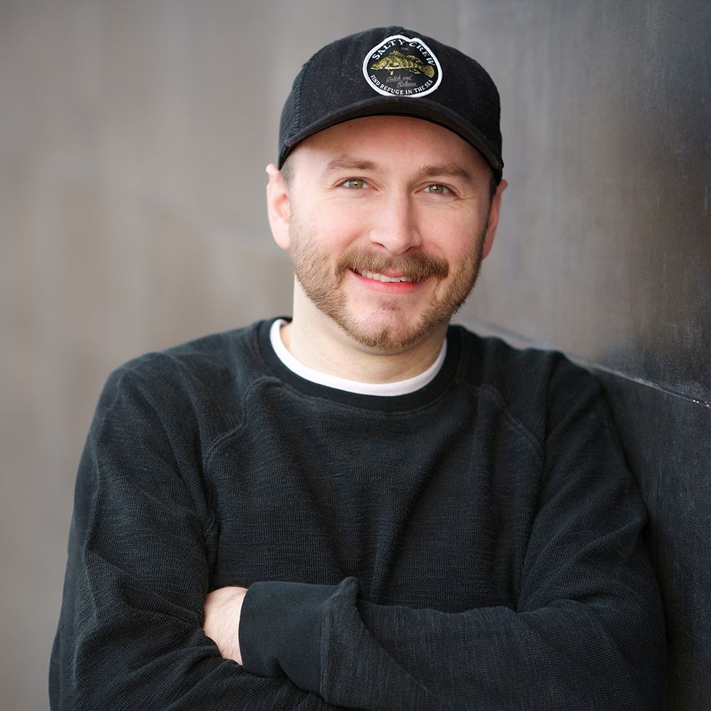 A man wearing a black hat and a black sweater is smiling with his arms crossed