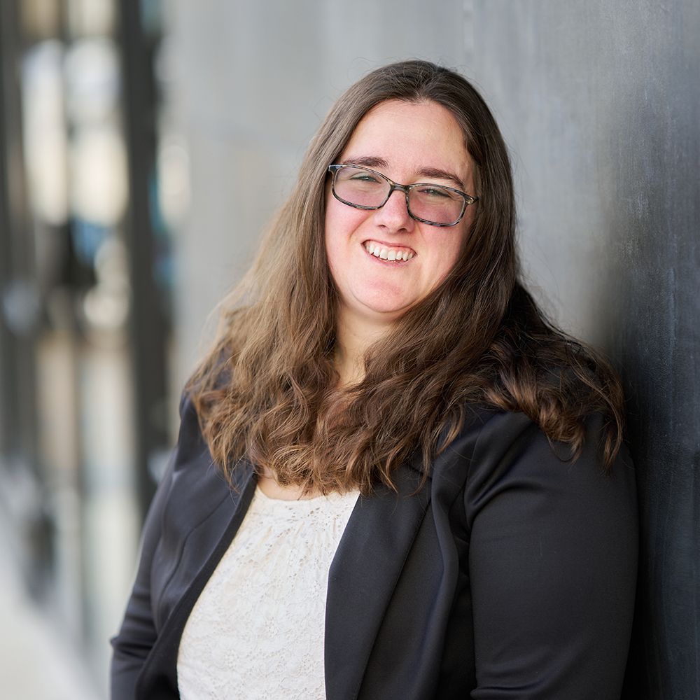 A woman wearing glasses and a black jacket is leaning against a wall and smiling.