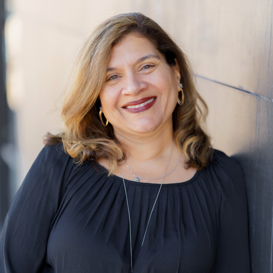 A woman in a black dress is smiling and leaning against a wall.