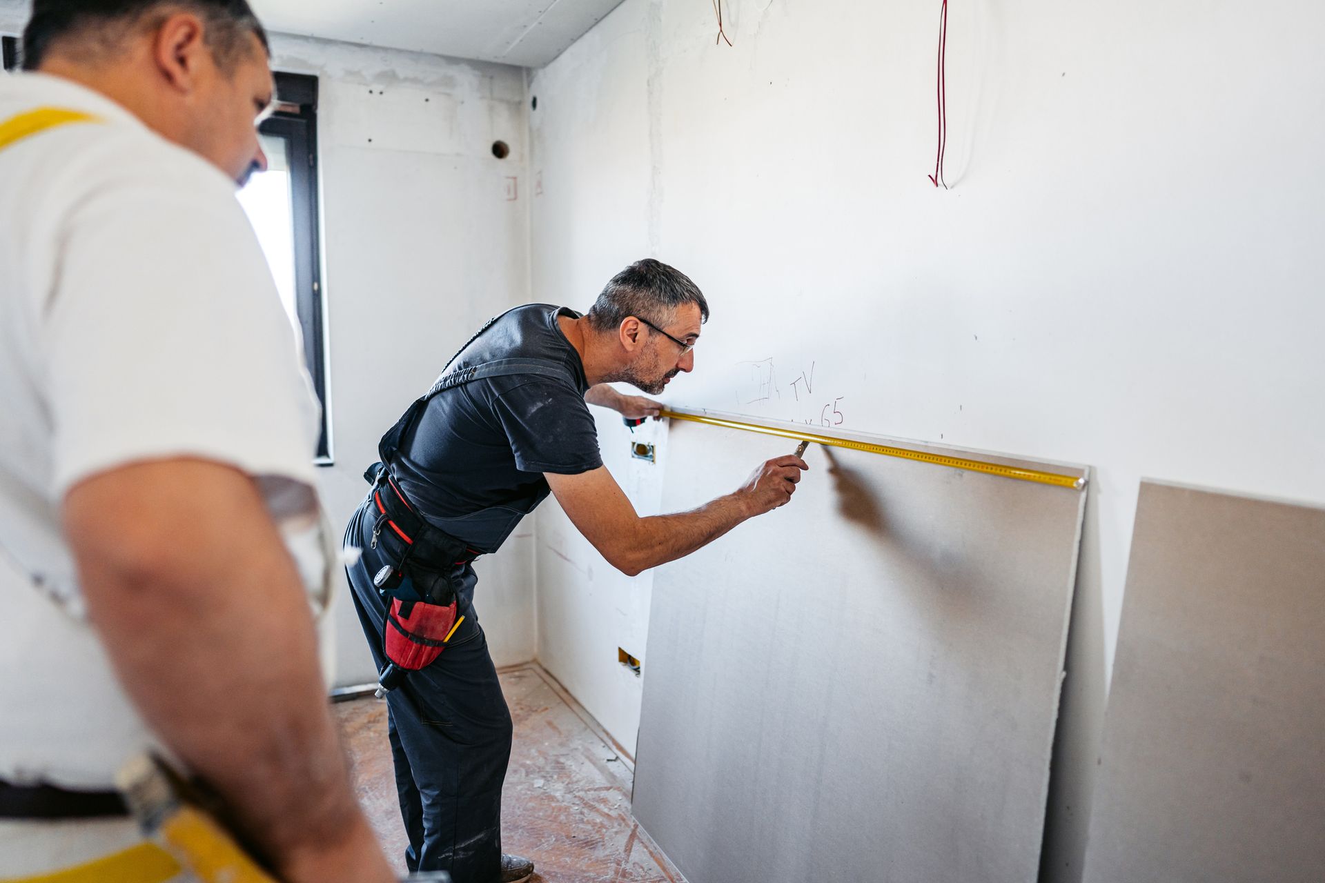 Two workers measuring wall space for drywall installation inside a room under construction.