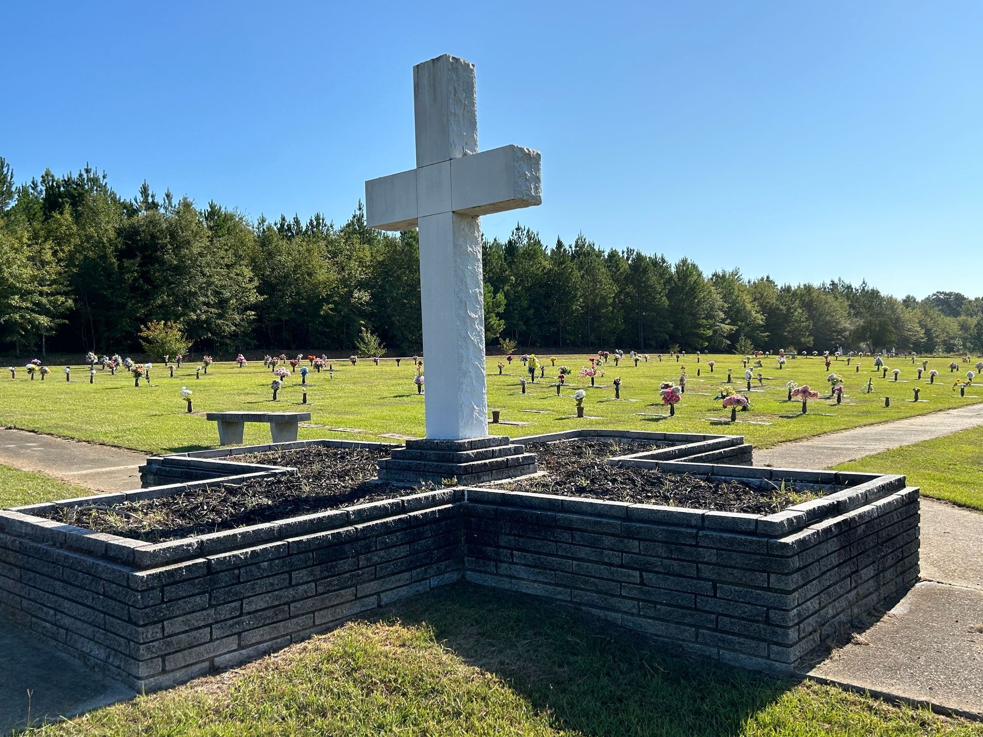 White cross monument in a cemetery with headstones and trees under a blue sky.
