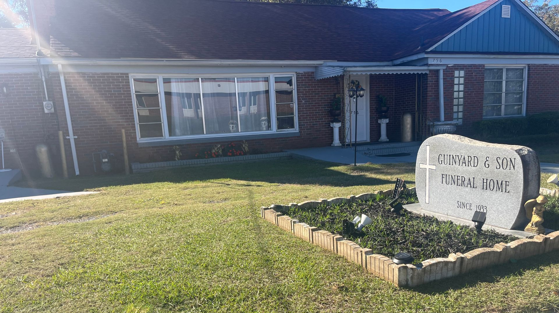 Exterior of a brick funeral home with a large stone sign in a flower bed, sunny day.