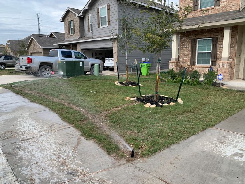 Sprinkler watering a green lawn in front of suburban houses; truck parked in the background.