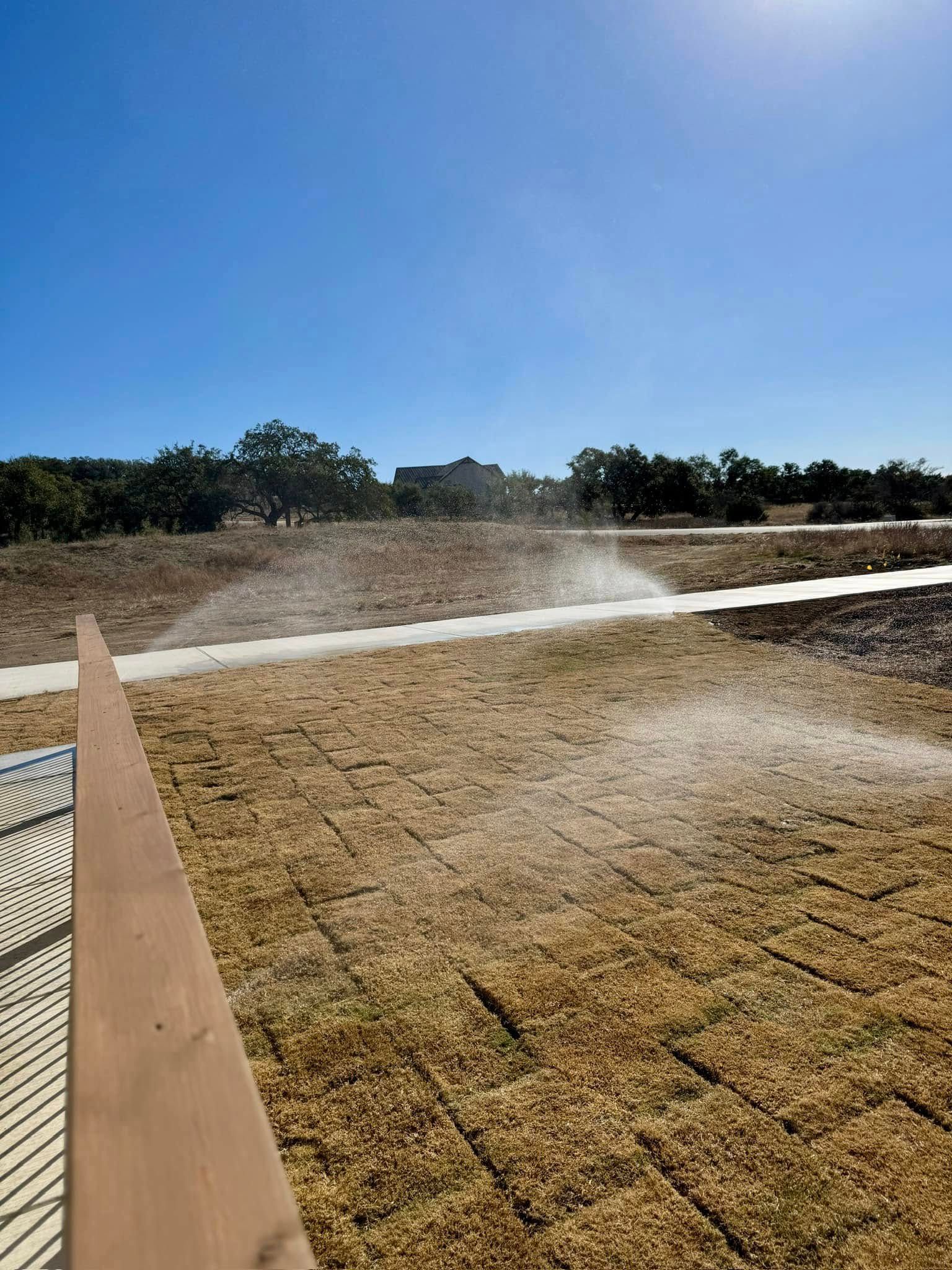 Sprinklers watering a brown, dry grassy area under a bright blue sky.
