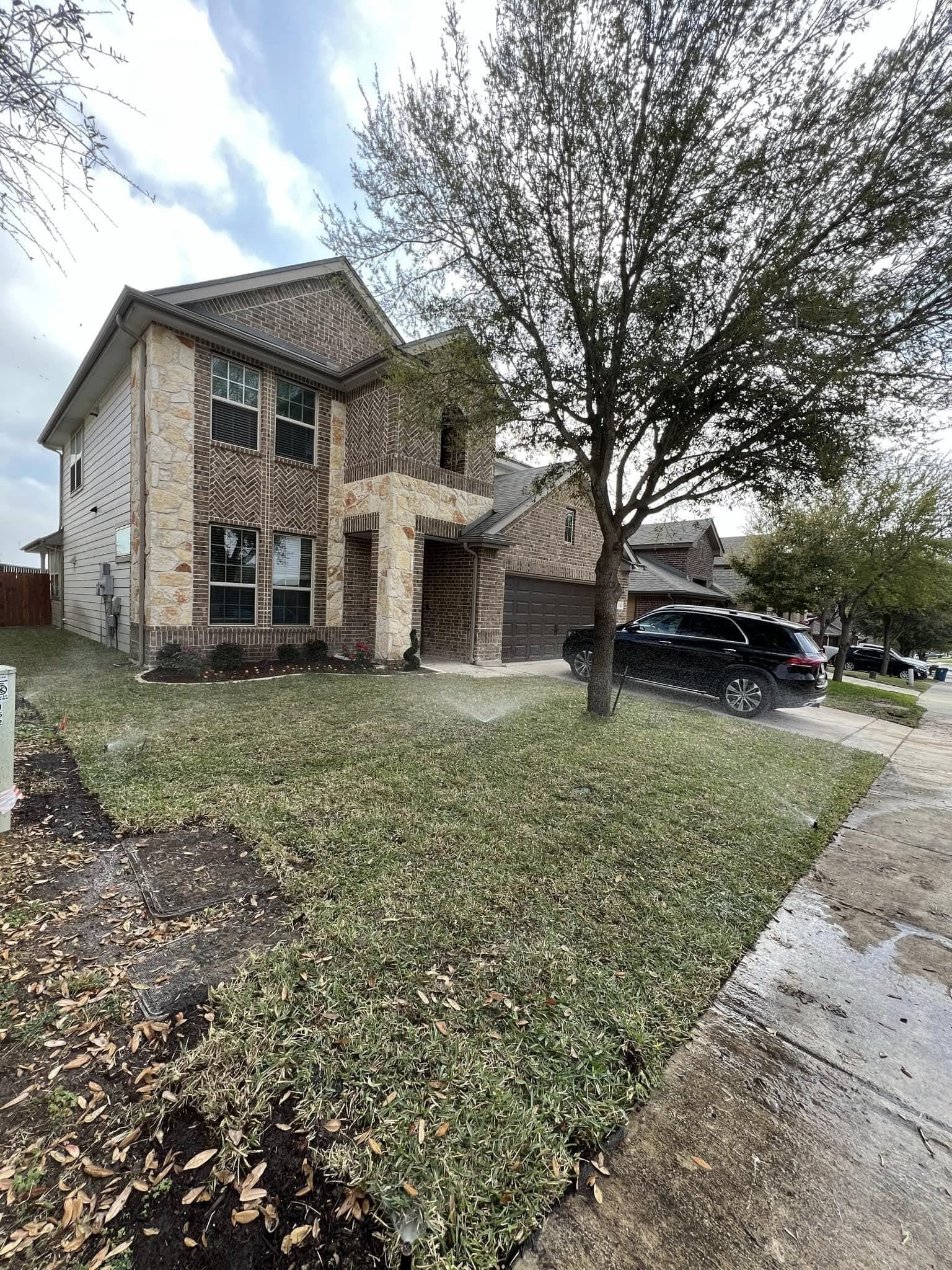 Two-story brick home with green lawn, tree, and car parked in driveway. Cloudy sky.