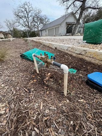 Sprinkler system components in a garden bed with mulch. Brown and green with a house in the background.