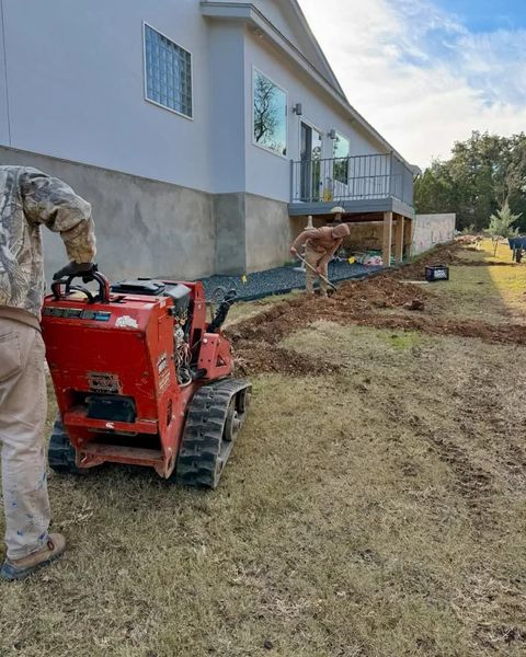 Workers operating a small red trencher next to a house, digging in the yard for landscaping.