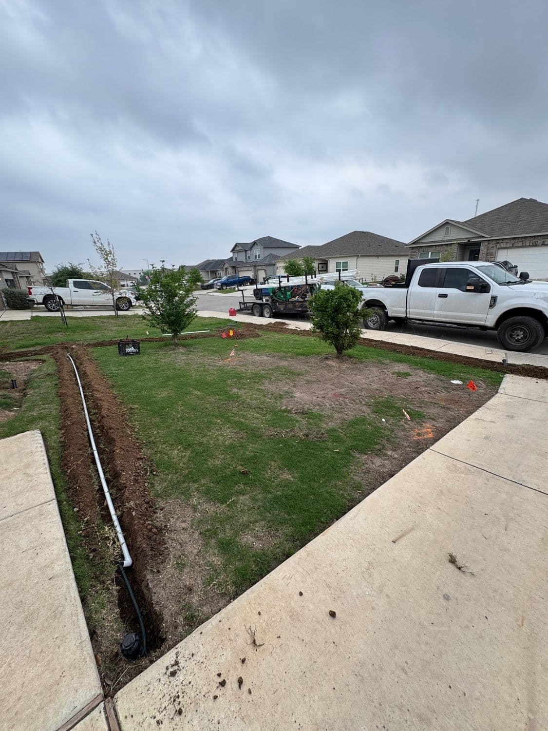 A lawn with a trench dug, white truck parked in front. Cloudy sky.