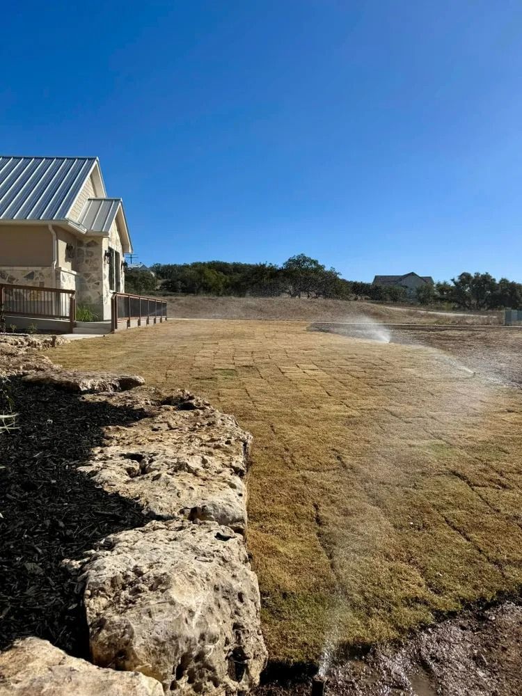 A house with a dry lawn being watered by a sprinkler system, near a rocky ledge under a blue sky.