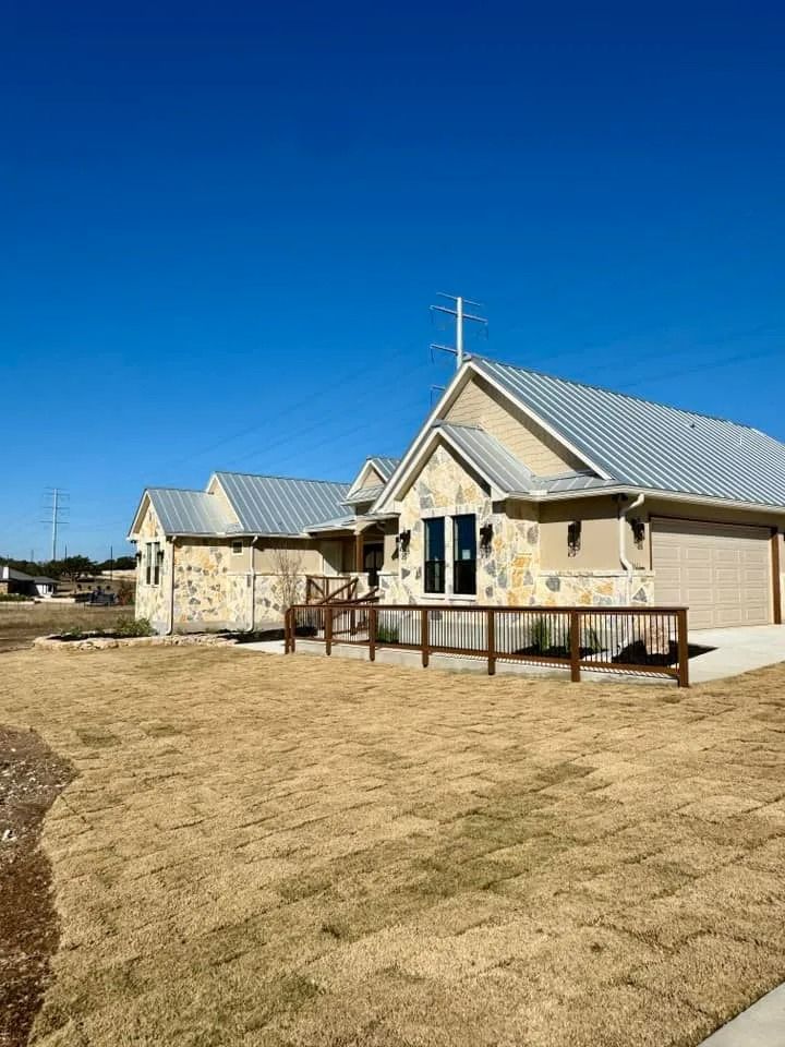 Stone house with metal roof, brown fence and dry grass under a blue sky.