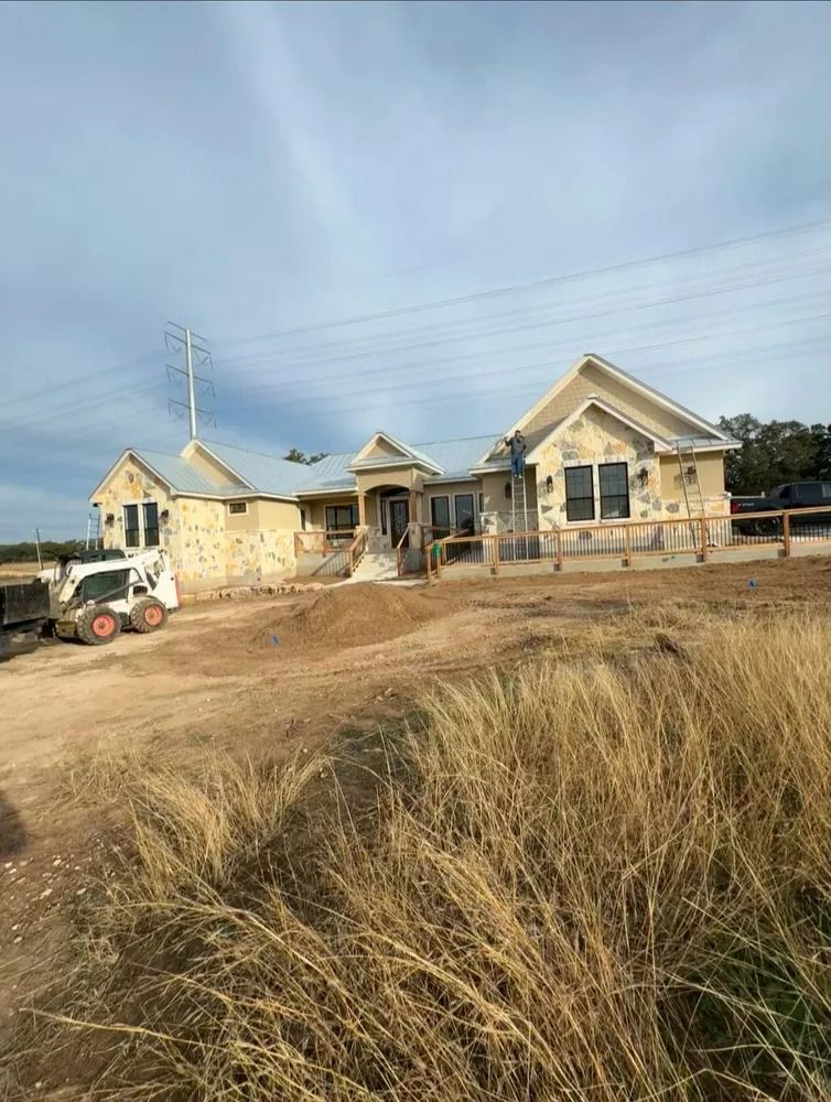 House under construction; light stucco, small dozer, dirt and tall grass, cloudy sky.