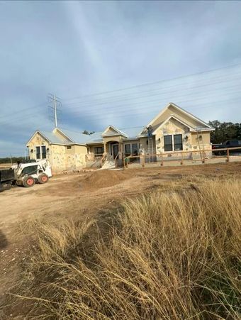House under construction; light stucco, small dozer, dirt and tall grass, cloudy sky.