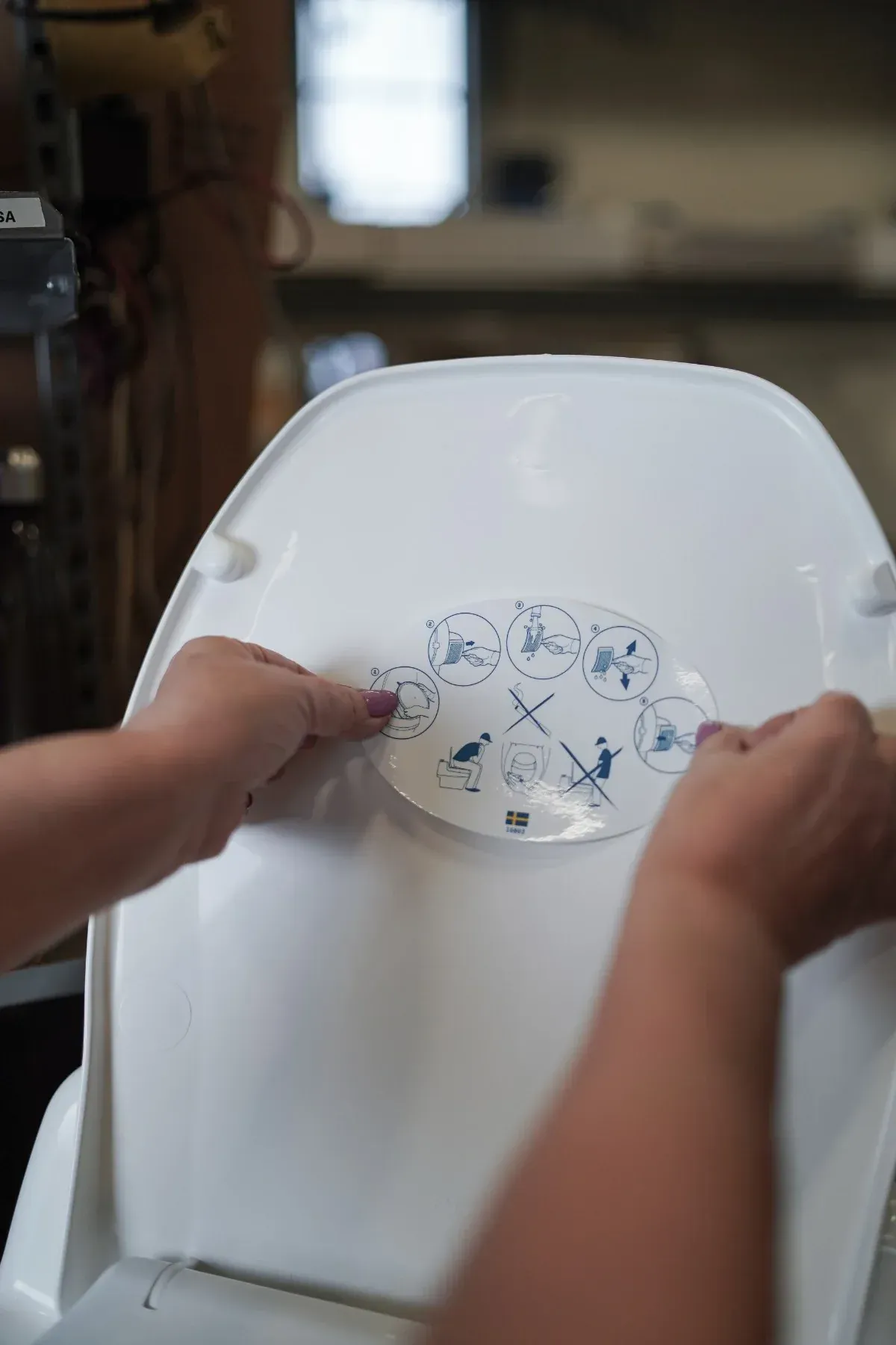 Person's hands attaching a safety label with blue icons to a white high chair back. Blurred workshop background.