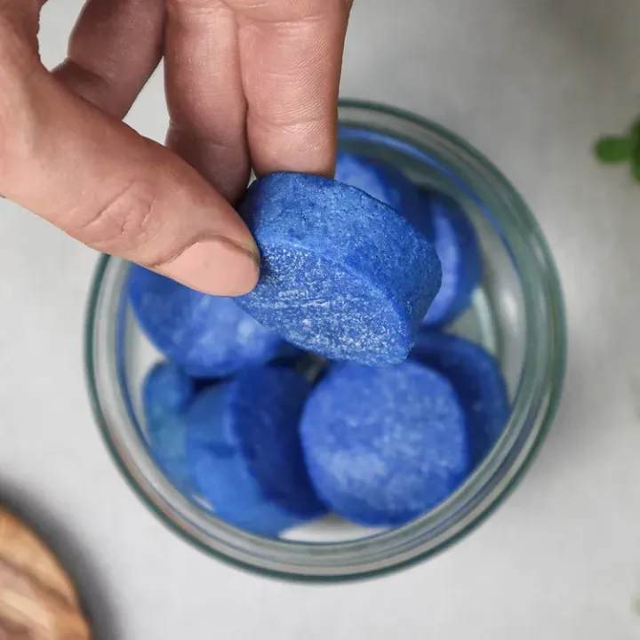 A hand holding a blue, disc-shaped item above a clear jar filled with more of the same.