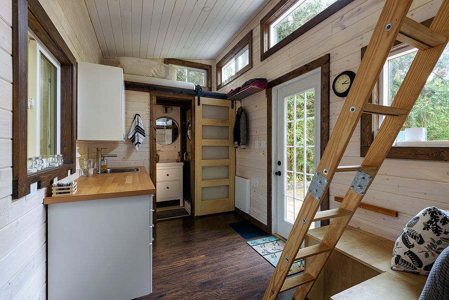 Interior of a tiny home with wood flooring, a white kitchen, and a ladder leading to a loft. A door leads to a bathroom.