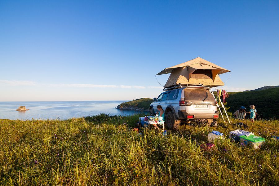 SUV with rooftop tent parked on a grassy cliff overlooking the sea. People gather around a table.