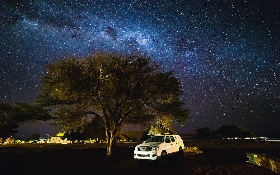 A white SUV parked under a tree, silhouetted against a vibrant night sky filled with stars and the Milky Way.