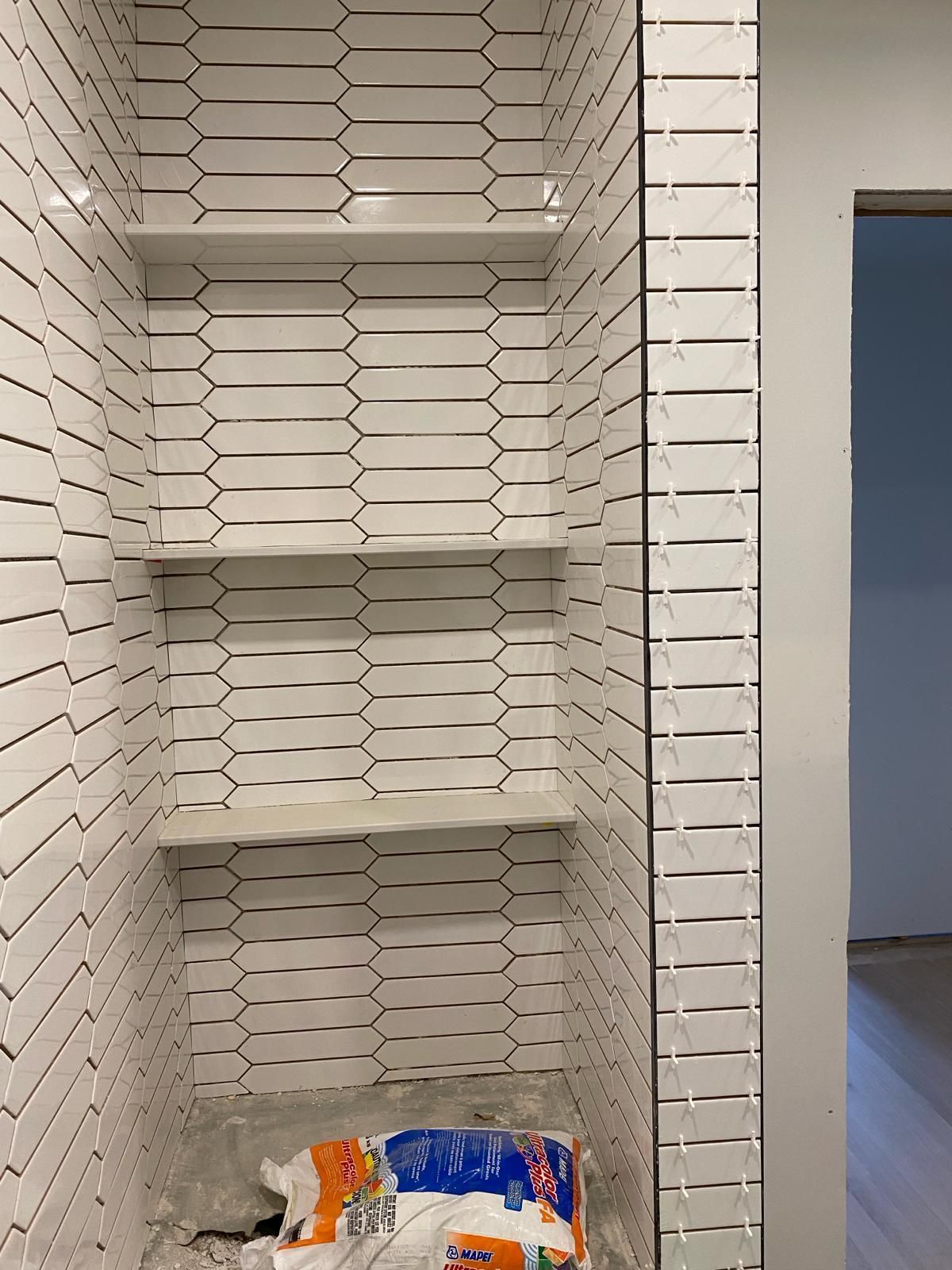 A bathroom with white tiles and shelves and a bag of toilet paper.