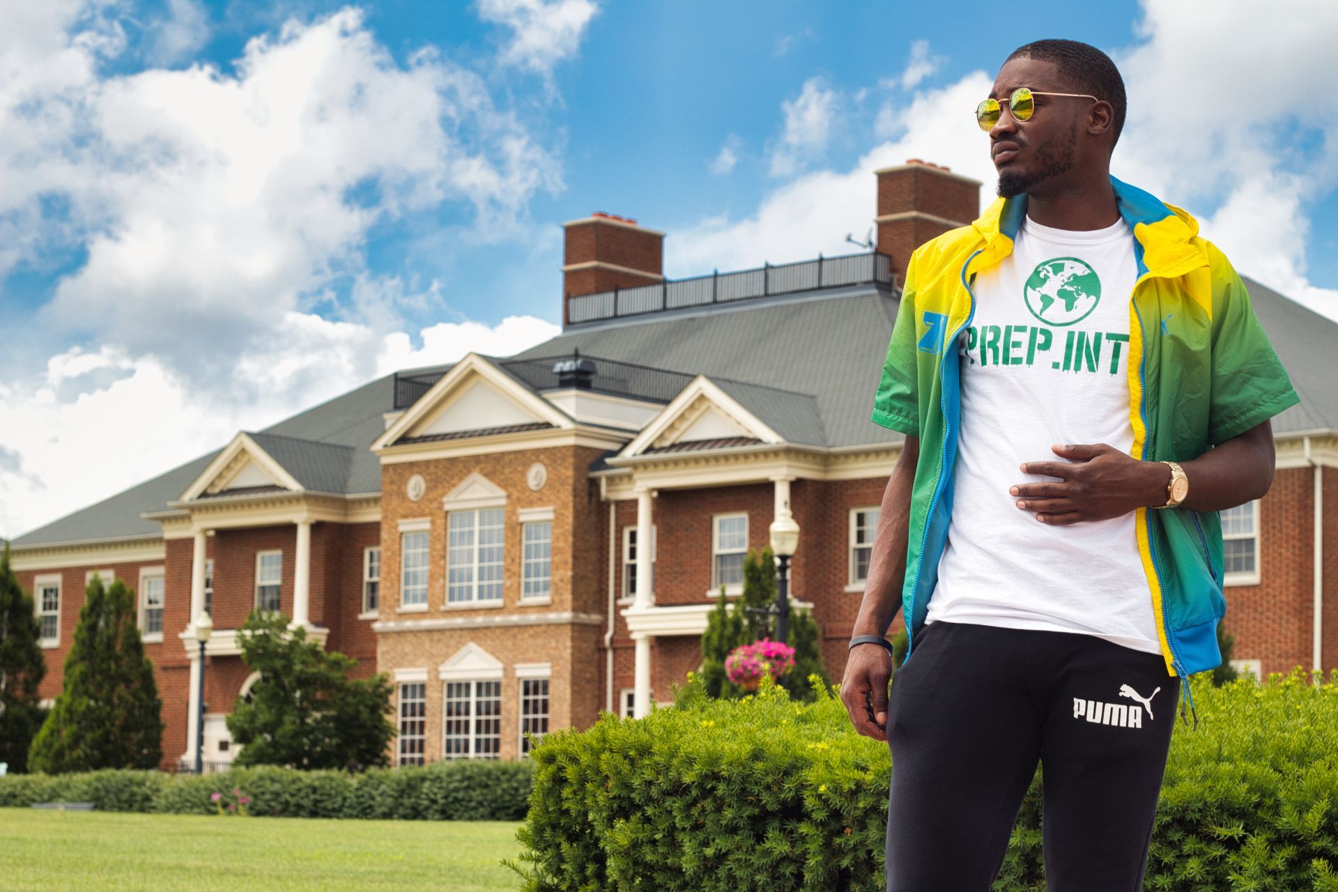 Man in sunglasses stands in front of a large brick house. He wears a white shirt, Puma pants, and a green/yellow jacket.