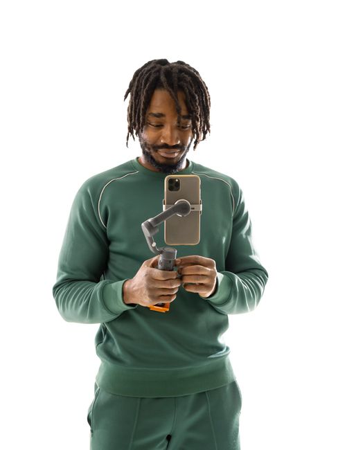 A man in a green sweatshirt is holding a gimbal and looking at his phone.