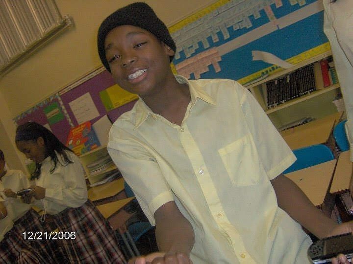 Teenager in yellow shirt and black beanie in a classroom, smiling, hands up.