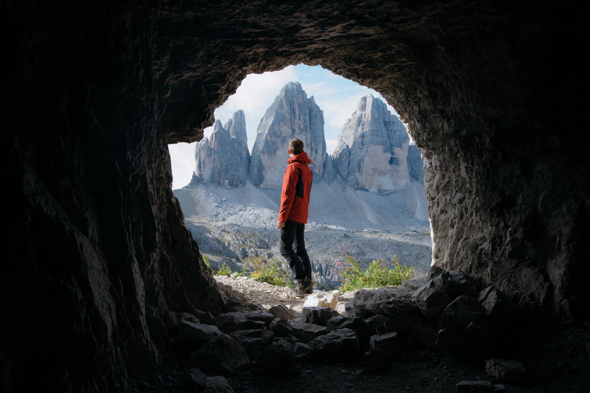 A man in a red jacket is standing in a cave looking out at a mountain range.