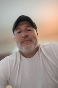 Man with glasses and grey beard in a blue shirt smiles in front of a bookshelf.