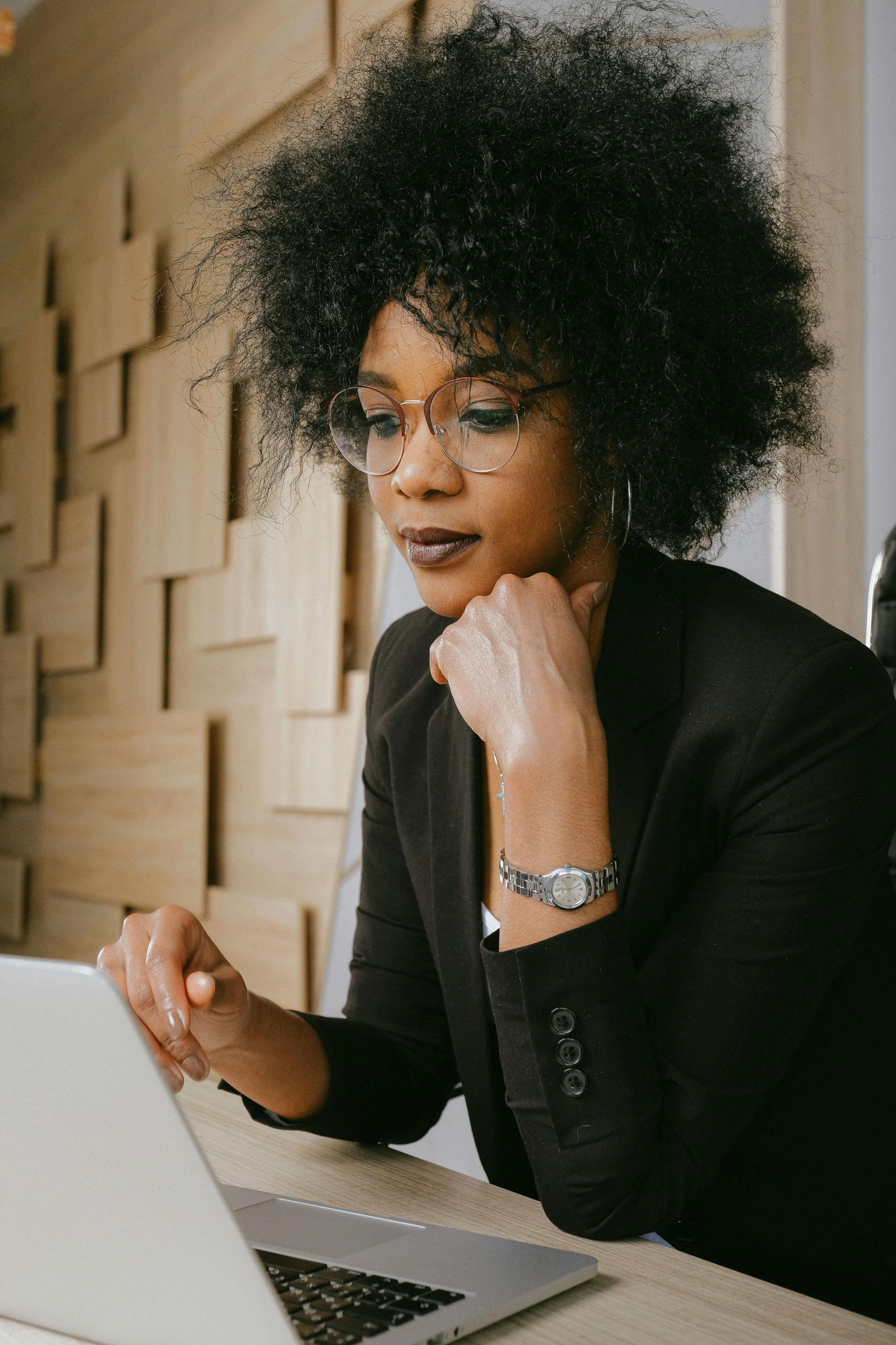 Woman with afro, glasses, and blazer, working on a laptop at a wooden desk, thoughtful expression.