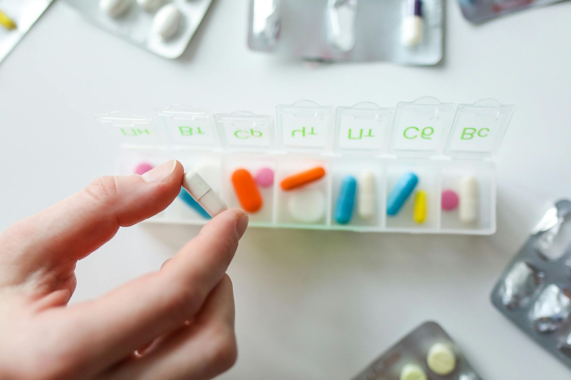 Hand holding a white and blue pill from a pill organizer filled with various colored pills, blister packs in background.