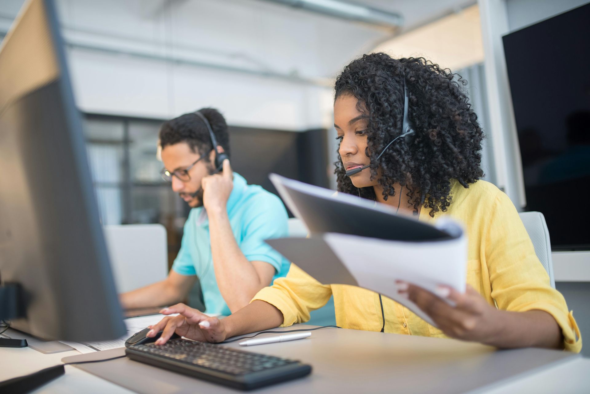 Two businesspeople analyze financial data, comparing a printed chart to a laptop screen, in a bright office setting.