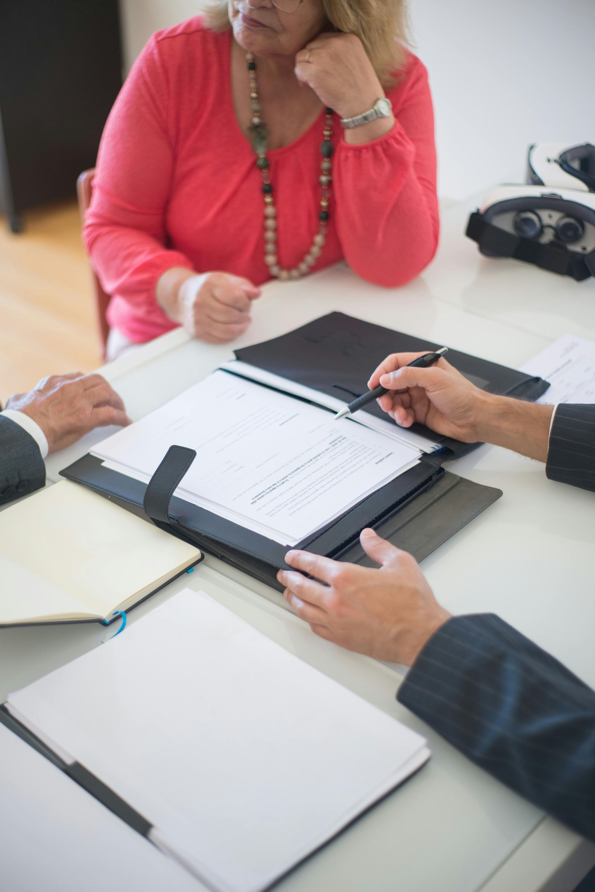 A woman in a pink shirt sits at a table while a person in a suit points to a document. Others sit nearby.