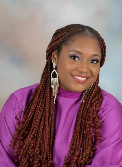 A woman with long, braided hair and large earrings smiles, wearing a purple top.