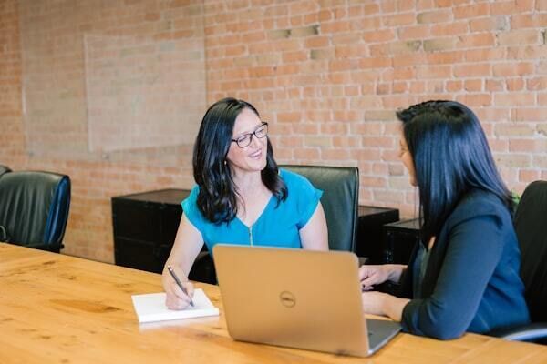 Two women at a wooden table, one writing and smiling, the other using a laptop in a brick-walled office setting.