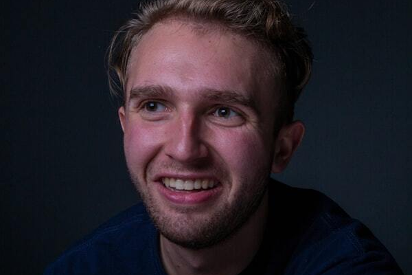 A young man with blond hair smiles broadly, looking to the side. He wears a dark blue shirt, set against a dark background.