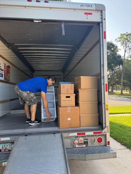 Man loads boxes and a plastic organizer into a moving truck. He wears a blue shirt and camo shorts.