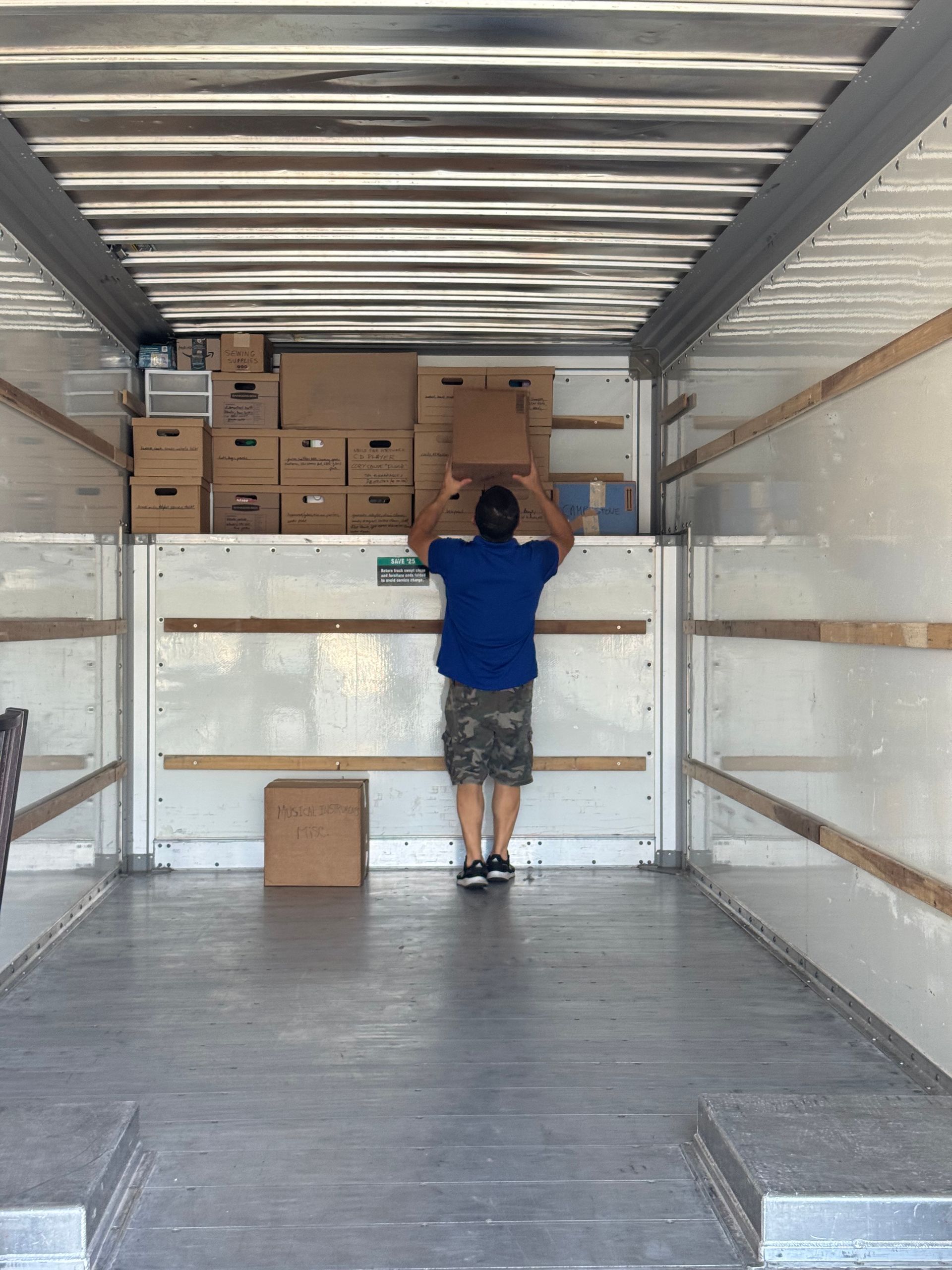 Man inside moving truck, loading boxes onto a shelf. White walls, grey floor, boxes stacked.