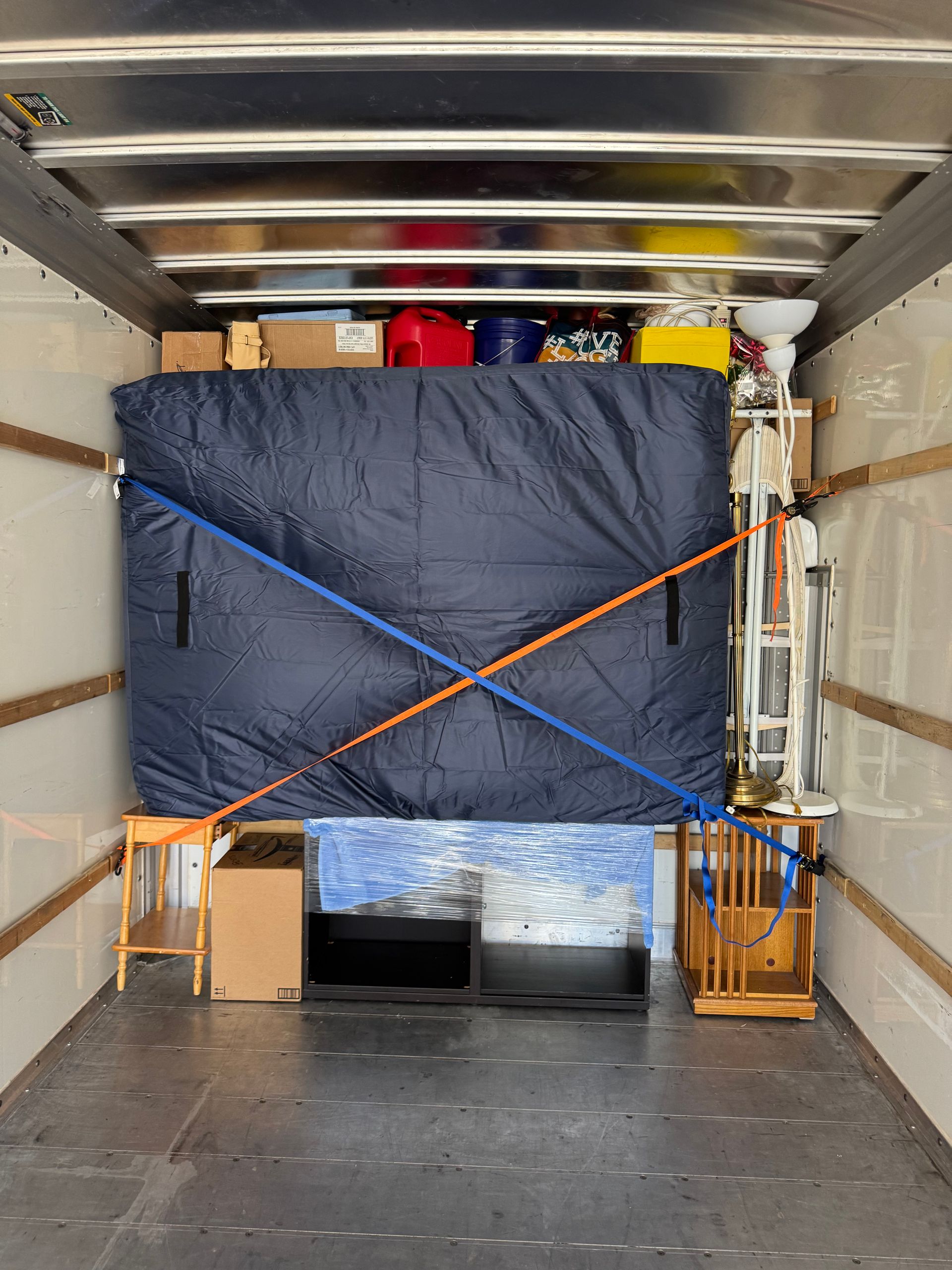 Interior of a moving truck, loaded with furniture and boxes. A mattress is strapped down, items are on shelves.