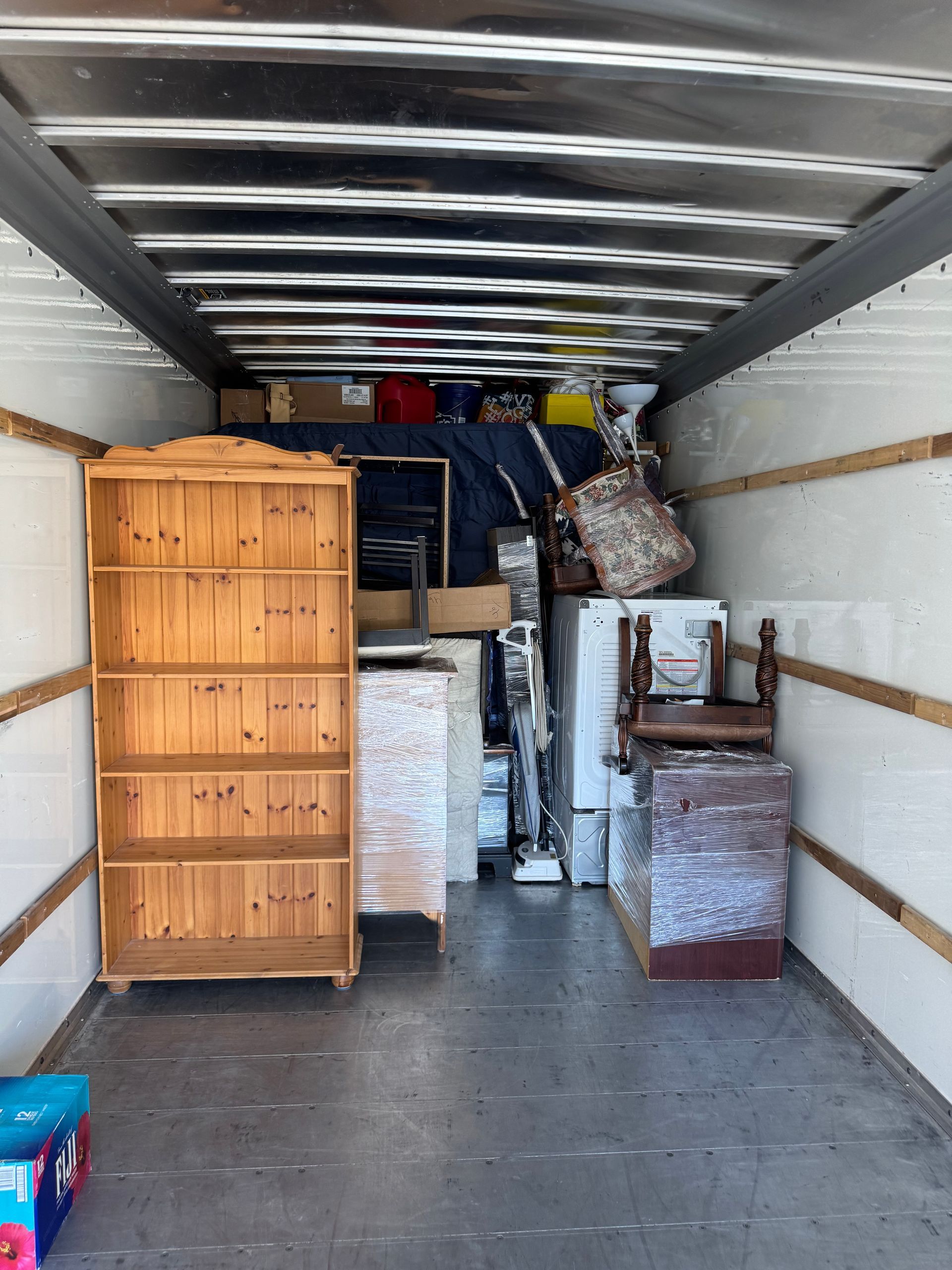 Interior of a moving truck filled with furniture, including a bookshelf, dresser, and wrapped items.