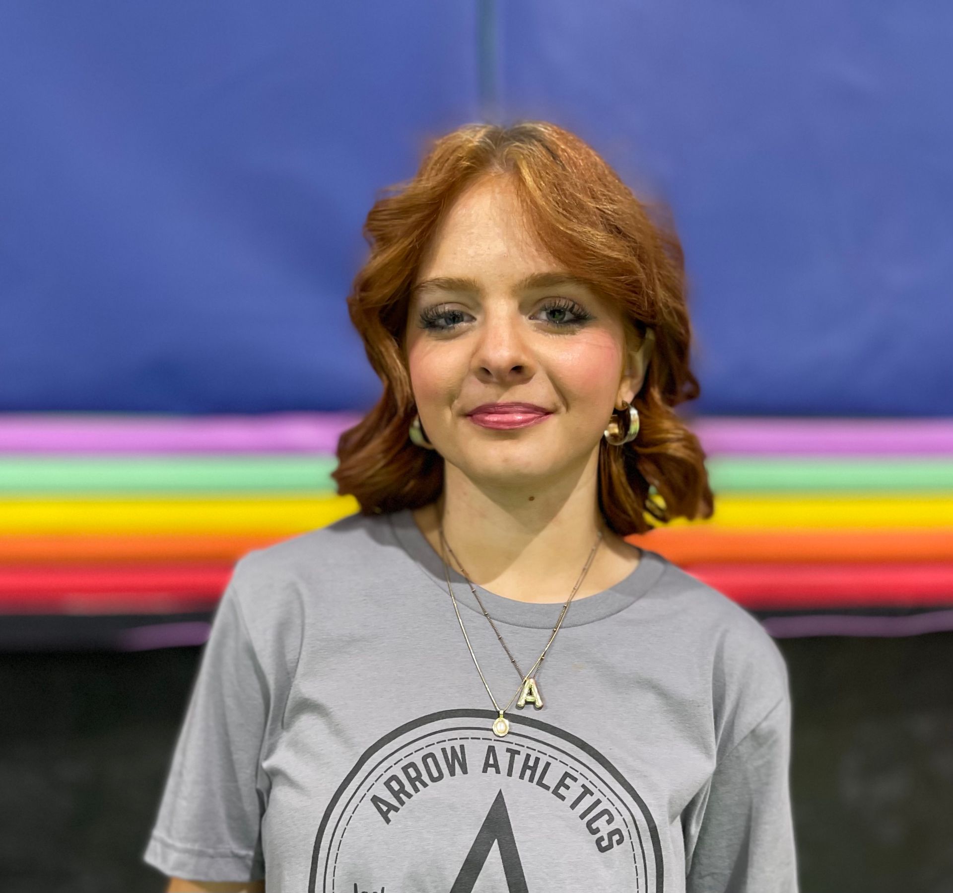 A woman wearing an arrow athletics shirt is standing in front of a rainbow colored wall.