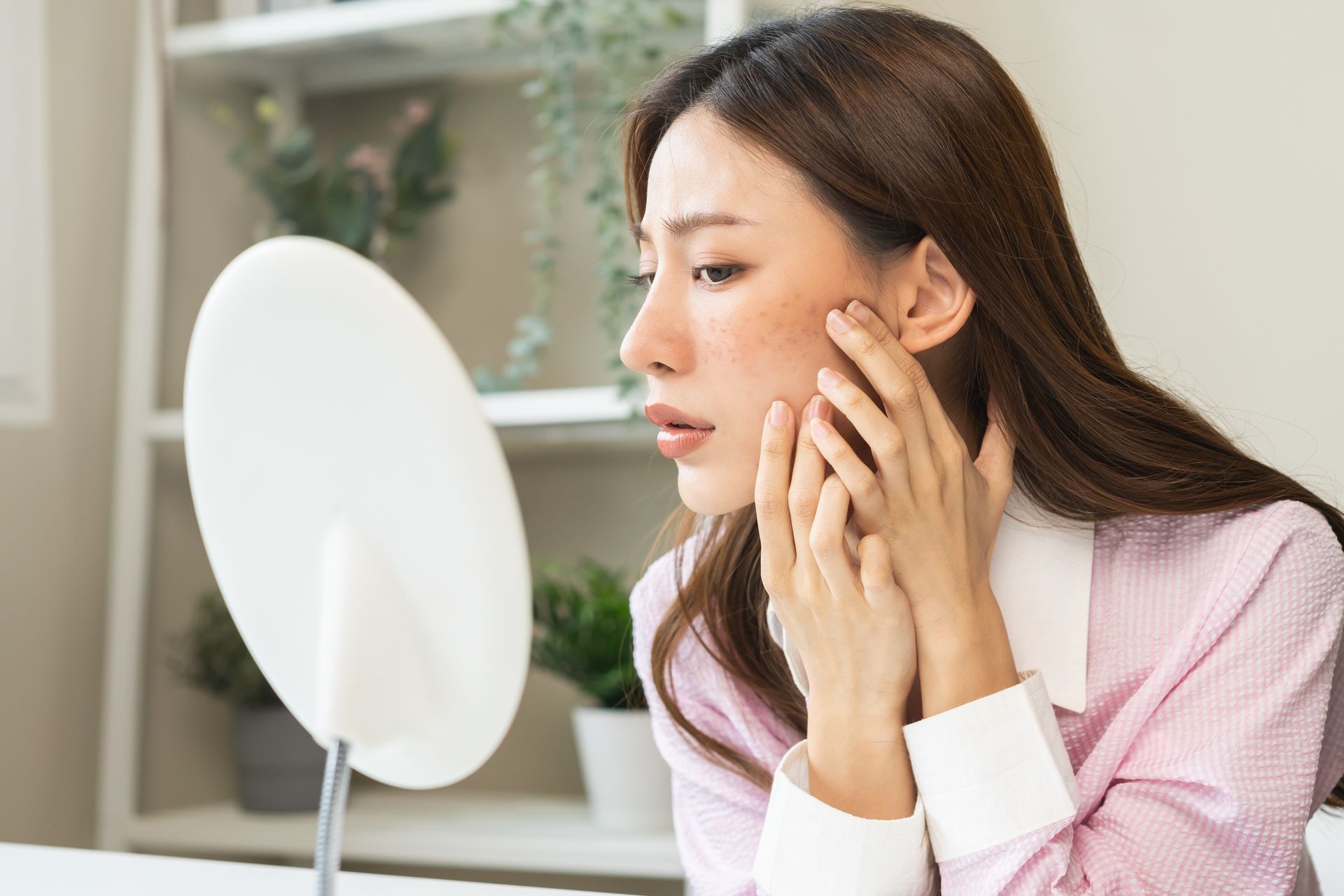 Woman looking at face in mirror, examining skin imperfections with a concerned expression.